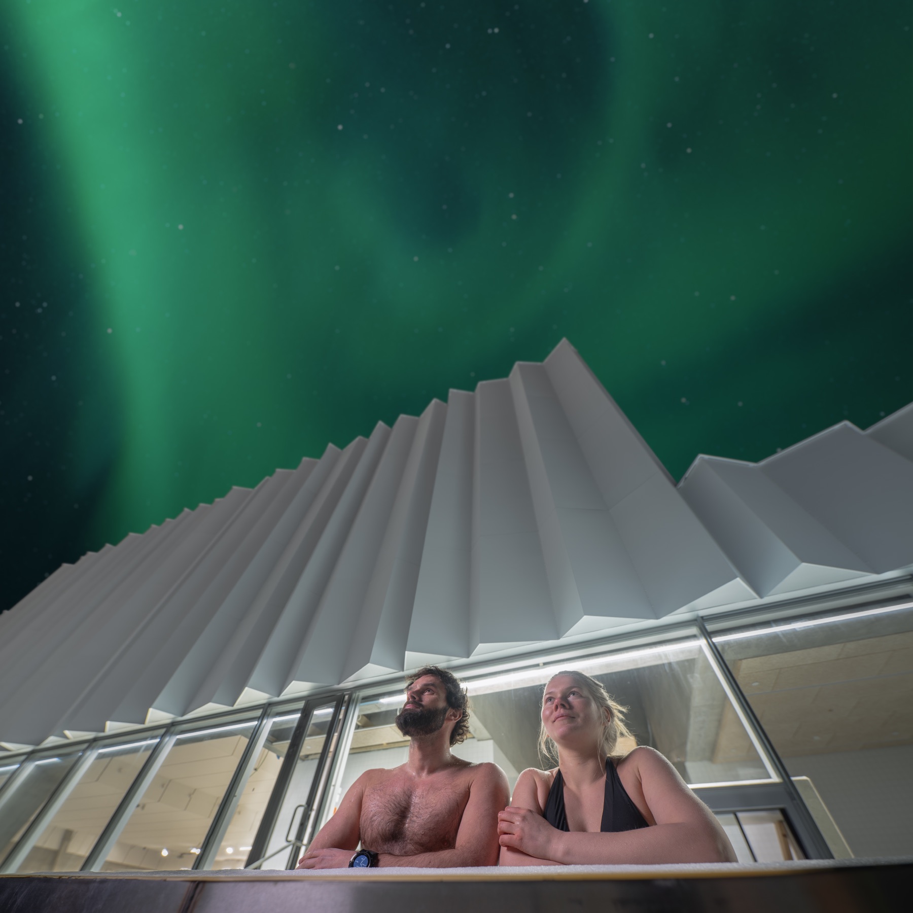 Two guests at the outdoor pool beneath the northern lights above Tromsøbadet.