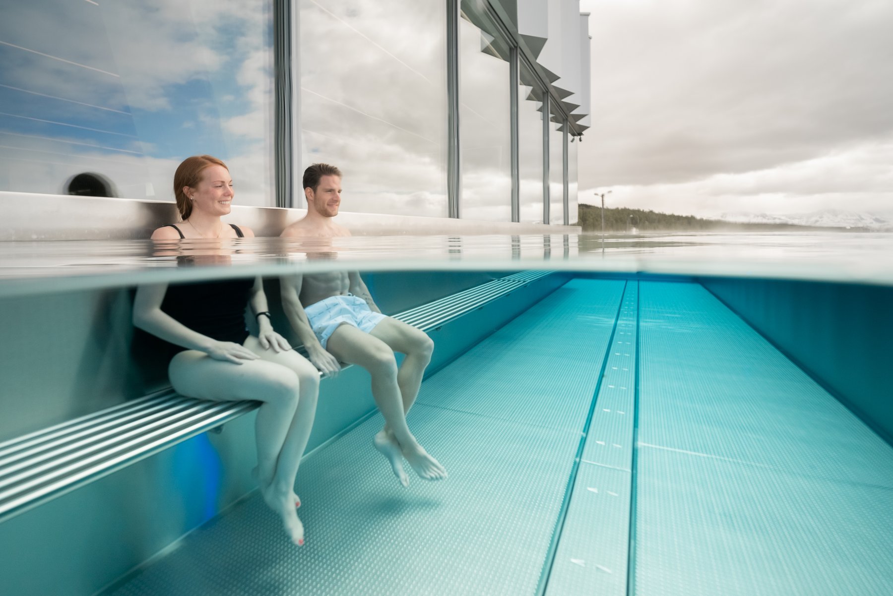 Two guests sitting on a submerged bench in the outdoor pool at Tromsøbadet, photographed above and below the water.